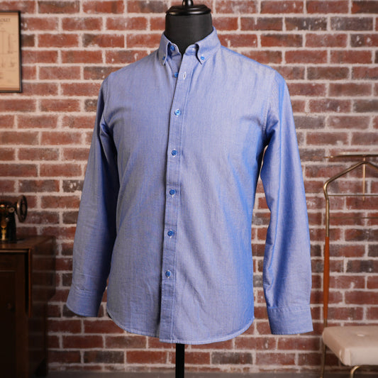 A blue long-sleeve oxford cloth button-down shirt displayed on a black mannequin torso, set against a red brick wall background with vintage decor on the left and a chair on the right.