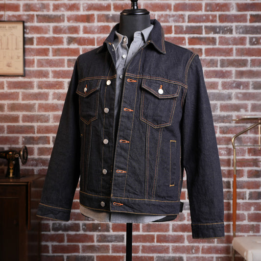 A dark indigo denim jacket with orange-brown contrast stitching, displayed on a black mannequin torso against a red brick wall background.
