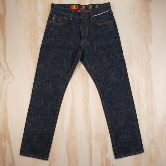 Dark indigo blue selvedge denim jeans laid flat on a light wood grain background, featuring a red and yellow waistband label and a red circular patch on the front pocket.