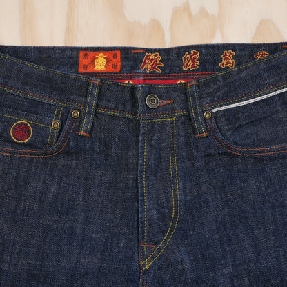 Close-up of dark indigo selvedge denim jeans laid flat on a light wood background, showing the waistband with red embroidered Chinese characters, gold-tone metal hardware, and contrast yellow stitching and selvedge ID detail above left pocket.
