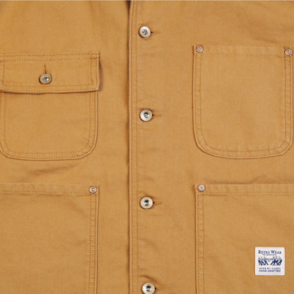 Close-up of a khaki canvas chore coat showing two front pockets with metal rivets and a white sewn-in label that reads 'RETRO WEAR MADE BY MADEN HAND-CRAFTED'.