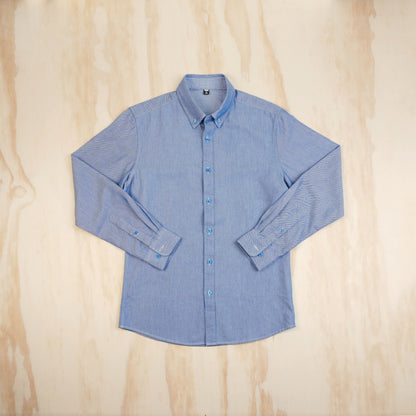 A light blue long-sleeve button-up oxford cloth shirt laid flat on a light wood background, viewed from the front.