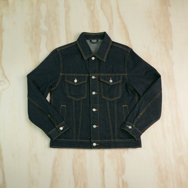 A dark indigo type 3 selvedge denim trucker jacket with contrast gold/yellow stitching, laid flat on a light wood grain background.