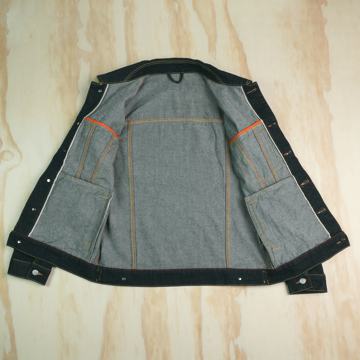 A Type 3 selvedge denim jacket laid flat on a light wood background, showing the interior side with gray denim fabric, selvedge ID trim on button plackets, internal dump pockets, orange contrast stitching, and metal rivets.