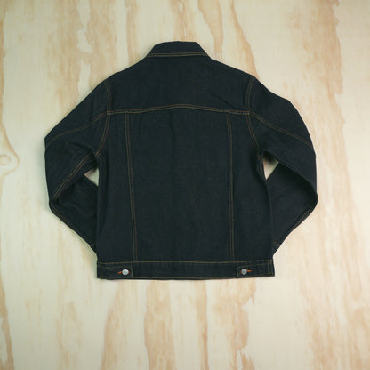 A dark indigo type 3 selvedge denim jacket shown from the back, laid flat on a light wood grain surface. The jacket features contrast orange stitching and silver metal snap buttons at the bottom hem.