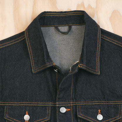 Close-up of a dark indigo type 3 selvedge denim jacket with orange contrast stitching, featuring a pointed collar, button front, and two buttoned chest pockets, laid flat on a light wood background.